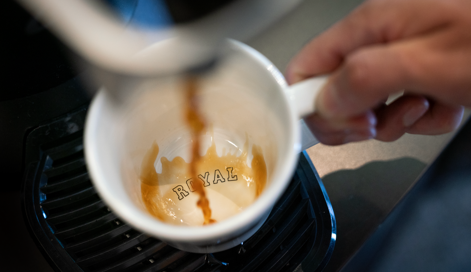 Person pouring coffee into a white mug with 'ROYAL' branding on a blurred background