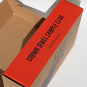 A brown cardboard sample box with a bright red top panel printed with black text reading 'CROWN JEWEL SAMPLE CLUB' and smaller 'ROYALDEFFICE.COM'.