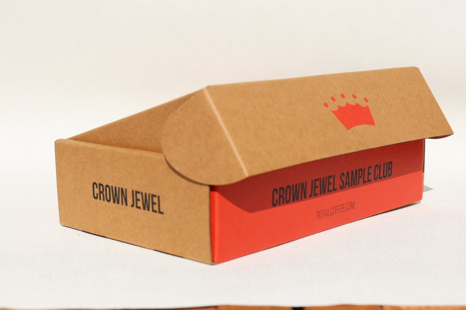 A brown cardboard sample box with a red side panel and flap, printed with the text 'CROWN JEWEL SAMPLE CLUB' and a small red crown logo. The box is photographed on a plain white background.